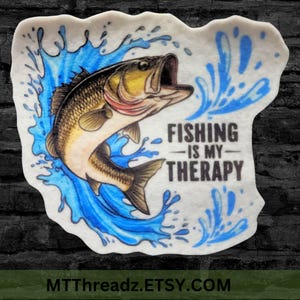 May include: A white sticker featuring a blue and green illustration of a bass fish leaping from the water. The text "FISHING IS MY THERAPY" is printed in black beneath the fish.