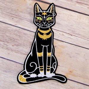 May include: Black cat patch with Egyptian-inspired design. The cat is black with gold and white accents, including a collar, leg bands, and facial details. The eyes are green. The patch is embroidered with a white border.