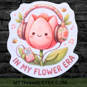 May include: A sticker featuring a pink tulip wearing floral headphones and holding a pink music player. The text "IN MY FLOWER ERA" is written in pink. Two cartoon bees and musical notes are also included.