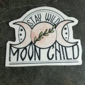 May include: Black and white sticker with a triple moon symbol and the text "Stay Wild Moon Child". The moon is pink and has a green leaf sprig inside.