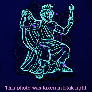 May include: A neon blue embroidered patch featuring a seated figure with a crown and scepter, illuminated under black light. The design includes constellation lines and the text "This photo was taken in blak light".