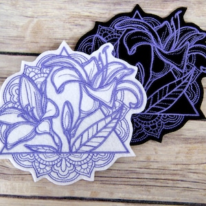 May include: Two embroidered patches featuring a stylized lily flower design. The patches are white and black with purple stitching. The design includes a mandala-like border.