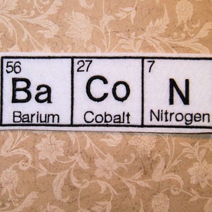 May include: White felt patch with three squares, each representing a chemical element. The first square shows the element Barium (Ba) with the atomic number 56. The second square shows the element Cobalt (Co) with the atomic number 27. The third square shows the element Nitrogen (N) with the atomic number 7.