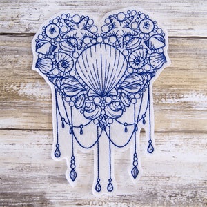 May include: A white felt heart-shaped patch with blue embroidery of seashells, starfish, and pearls. The design features a large seashell in the center and a dangling chain of pearls and beads.