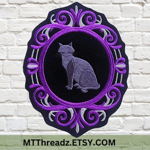 May include: An embroidered patch featuring a gray cat sitting in profile within a black circular center. The cat is surrounded by a decorative frame of purple and silver scrollwork embroidery on a black background with a scalloped edge.