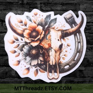 May include: A sticker featuring a watercolor design of a bull skull with large horns, adorned with flowers and foliage in shades of orange, brown, and gray. A horseshoe is positioned to the right. The text "MTThreadz.ETSY.COM" is at the bottom.