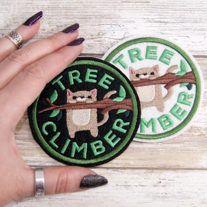 May include: Two embroidered patches with a cat hanging from a branch. The patches are round and have green and white borders. The text on the patches reads "Tree Climber".