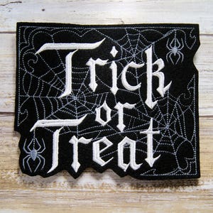 May include: Black felt patch with white embroidered text "Trick or Treat" in a gothic font. The patch is decorated with a spider web design and two small spiders.