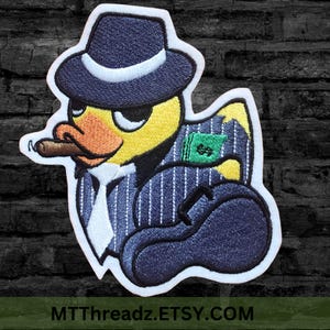 May include: Embroidered iron-on patch of a yellow duck wearing a blue pinstripe suit, a fedora hat, and holding a cigar and money. The patch has a white outline.