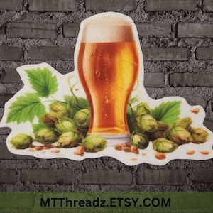 May include: A glass of beer with a white head, surrounded by green hops. The image has the text "MTTThreadz.ETSY.COM" at the bottom.