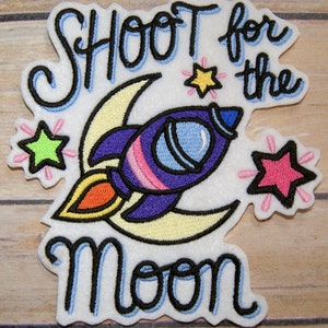 May include: Embroidered iron-on patch with a purple rocket ship flying past a yellow crescent moon. The text "Shoot for the Moon" is in blue and black lettering. There are three stars in different colors.