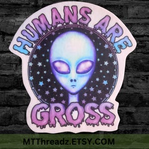 May include: A sticker featuring an alien with large purple eyes and a light blue face, set against a starry background. The words "HUMANS ARE GROSS" are written in a dripping font around the alien's head. The sticker has a white border.