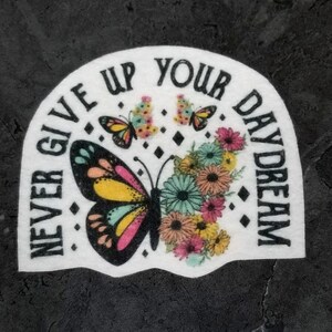 May include: White iron-on patch with a black outline and the text "Never Give Up Your Daydream". The patch features a large colorful butterfly with pink, yellow, and blue wings, surrounded by a cluster of pink, yellow, and blue flowers.