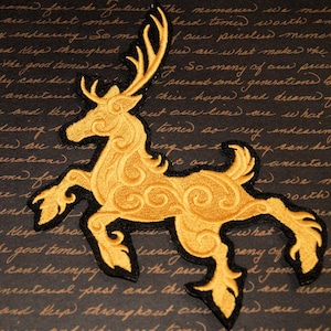 May include: A gold embroidered patch of a stylized deer with intricate swirling details. The deer is facing left and has large antlers. The patch is on a dark brown background.