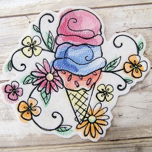 May include: Embroidered patch featuring a colourful ice cream cone with three scoops of ice cream and a waffle cone. The ice cream is pink, blue, and orange. The patch is surrounded by a floral design with green leaves and flowers in various colours.