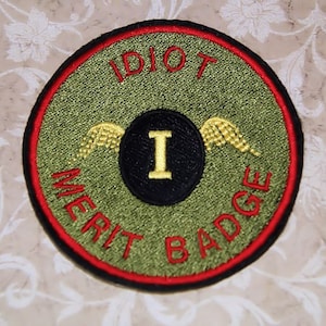 May include: A green and red embroidered patch with the text "Idiot Merit Badge" and a black circle with a gold "I" and gold wings.