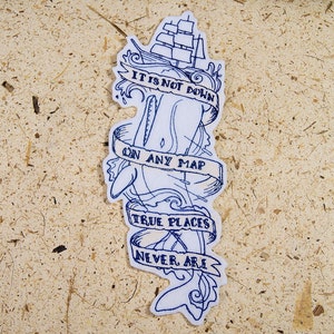 May include: White embroidered patch with a blue whale and a ship sailing on its back. The patch has a white ribbon with the text "It is not down on any map true places never are."