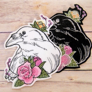 May include: Two embroidered patches featuring a black and white raven with floral accents. The white raven patch has a pink rose and green leaves. The black raven patch has a pink flower and green leaves.