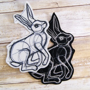 May include: Two embroidered felt patches of a rabbit, one in black and one in white. The black rabbit is on top of the white rabbit. Both rabbits are in a sitting position with their ears up.