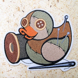 May include: Embroidered patch in the shape of a duck, featuring a patchwork design in shades of green, brown, and tan. The duck has button eyes, an orange beak, and a spool of thread. Pins and a needle are also included in the design.