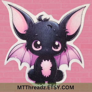May include: A black and purple cartoon bat with large eyes and pink ears. The bat is sitting on a pink background.