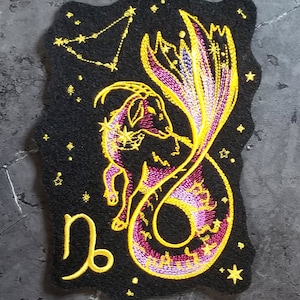 May include: Embroidered patch featuring a black and gold Capricorn zodiac sign with a pink and gold fish tail. The patch is on a black felt background with gold stars.