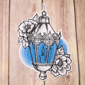 May include: A black and white embroidered patch of a lantern with a lit candle inside. The lantern is surrounded by two white flowers with blue accents. The background is a blue wash.
