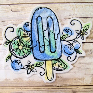May include: Embroidered patch featuring a blue popsicle with lime slices and blueberries. The design is surrounded by green leaves and white flowers.