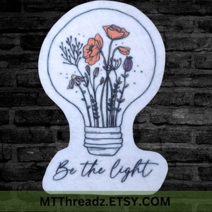 May include: A white light bulb with a black outline filled with orange and yellow flowers and green leaves. The text "Be the light" is written below the light bulb.