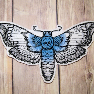 May include: A black and white embroidered patch of a blue and white moth with a skull in the center. The moth has intricate details and a gradient of blue to white on the wings.