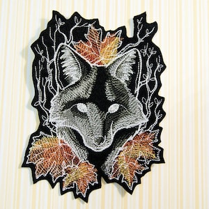 May include: Embroidered iron-on patch featuring a black and white fox head surrounded by autumn leaves. The fox has a white muzzle and white eyes. The leaves are in shades of orange, brown, and gold.