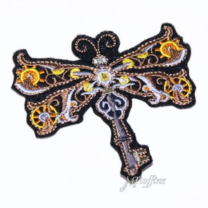 May include: A brown and gold embroidered iron key shaped like a dragonfly with intricate details. The key has a silver finish and is surrounded by a black fabric background.
