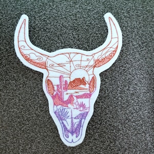 May include: Embroidered patch of a bull skull with a desert landscape inside. The skull is outlined in red thread, and the landscape is filled with shades of red, orange, and purple.