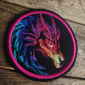 May include: A round embroidered patch with a black background and a colorful dragon design. The dragon is outlined in pink and has a rainbow-colored design.