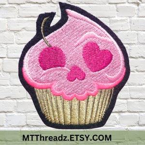 May include: Pink cupcake with a skull face and pink hearts for eyes. The cupcake has a cherry on top and a gold cupcake liner.