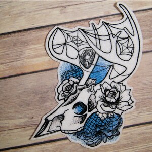 May include: Embroidered patch featuring a white deer skull with antlers, surrounded by blue and black roses and a blue spiderweb design.