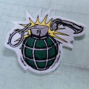 May include: An embroidered patch depicting a green and silver grenade with a yellow explosion behind it. The patch features the text "MTcoffinz.com" at the bottom.