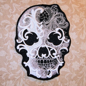 May include: A black and white embroidered skull patch with intricate floral designs. The skull is outlined in black and the floral designs are in white.