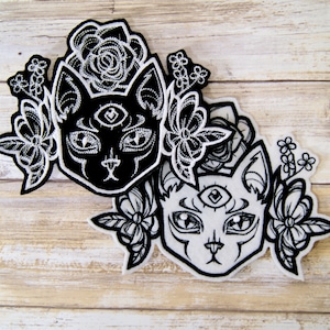 May include: Two black and white embroidered iron-on patches featuring stylized cats with a third eye and floral designs.