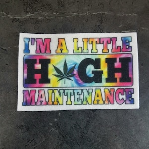 Puede incluir: Un parche rectangular con el texto "I'M A LITTLE HIGH MAINTENANCE" en letras coloridas de estilo tie-dye. La palabra "HIGH" presenta un gráfico de hoja de cannabis.
