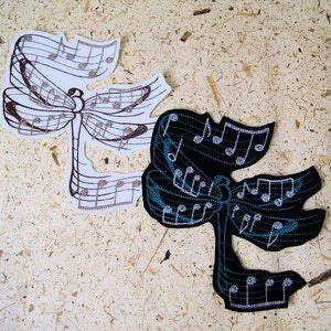 May include: Two embroidered patches in the shape of a stylized musical note. One is white with brown stitching, the other is black with teal and white stitching. The patches feature musical notes and lines.