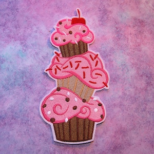Peut inclure: Une pile de trois cupcakes roses et bruns avec des pépites blanches et brunes. Le cupcake du dessus a une cerise rouge sur le dessus.