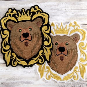 May include: Two embroidered iron-on patches featuring a brown bear wearing a gold crown, surrounded by a gold ornate frame. The patches are on a white wooden background.