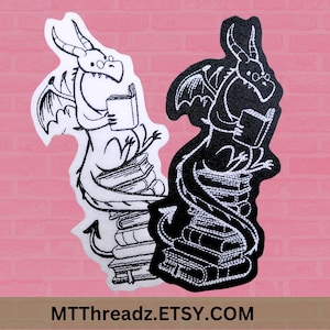 May include: Black and white embroidered iron-on patch of a dragon reading a book while sitting on a stack of books. The dragon's tail wraps around the books. The text "MTTThreadz.ETSY.COM" is at the bottom of the image.