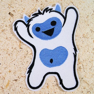 May include: White embroidered patch of a cartoon yeti with blue eyes, a black mouth, and a blue heart-shaped belly. The yeti is waving with its right arm.