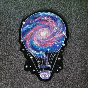 May include: Embroidered patch featuring a hot air balloon design with a galaxy theme. The balloon is filled with swirling blues, pinks, and purples, with white stars and a black border. The basket is purple and white.