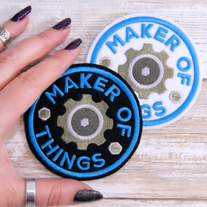 May include: Two embroidered patches with a blue border and a gear design. The top patch is white with the text "MAKER OF THINGS" in blue. The bottom patch is black with the text "MAKER OF THINGS" in blue.