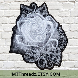 May include: Embroidered patch featuring a white rose design on a black background. The rose is intricately detailed with white thread, surrounded by a decorative border. The patch has a floral theme and is suitable for clothing or accessories.