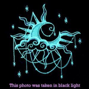 May include: A glowing blue embroidered design of a sun, moon, and clouds with stars on a black background. The design is reminiscent of a celestial scene.
