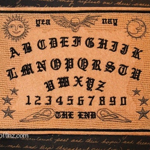 May include: A brown felt Ouija board with black embroidered letters, numbers, and symbols. The board has a skull and crossbones design at the top and the words "yea" and "nay" on either side of the skull. The board also has a crescent moon and a sun at the top corners. The words "THE END" are at the bottom of the board.